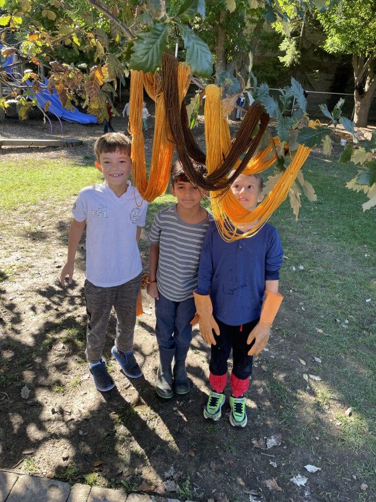 Three Da Vinci Waldorf School Third Graders Stand Beneath Skeins Of Golden Yellow, Orange, And Brown Naturally Dyed Wool Yarn Hanging From A Tree.